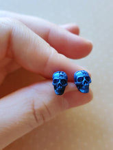 Load image into Gallery viewer, A close of picture of metallic  sapphire colored skull earrings held between finger and thumb.