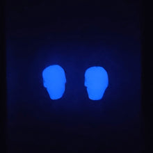 Load image into Gallery viewer, A piture of the skull earrings glowing in the dark. True color glows purplish.