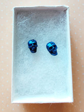 Load image into Gallery viewer, A close of picture of metallic  sapphire colored skull earring in a white paper jewelry box.