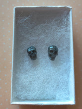 Load image into Gallery viewer, Grey colored skull stud earrings that shimmer silvery in the light inside a white paper jewelry box.