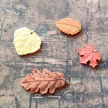 Load image into Gallery viewer, A picture showing a group of fall colored leaves for an example of various colors that the leaves could be customized to look. The group shows a red maple leaf, a rust orange beach leaf, a yellow aspen leaf, and a rust brown oak leaf.