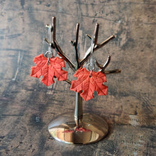 Load image into Gallery viewer, A pair of red maple leaf shaped polymer clay earrings are displayed hanging from a small, polished silver tree shaped display.