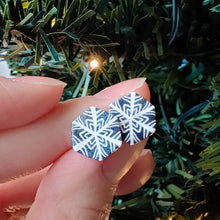 Load image into Gallery viewer, Two hexagon shaped earrings with a snowflake pattern in silver and white. The earrings are held between finger and thumb next to an artificial Christmas tree branch.