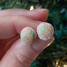 Load image into Gallery viewer, A pair of earrings made to look like sugar cookies decorated with green and red sugar sprinkles. The earrings are held between finger and thumb in front of an artificial Christmas tree.
