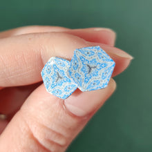 Load image into Gallery viewer, Two hexagon shaped earrings with intricate white, silver and blue patterns held between finger and thumb in bright sunlight.