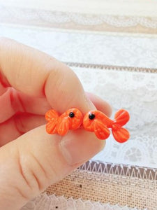 A pair of orange fish stud earrings held between finger and thumb