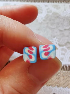 Two white, pink, and blue stud earrings held between finger and thumb