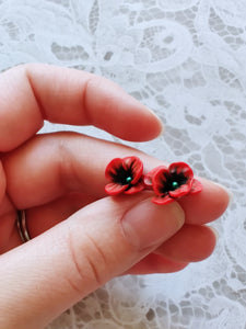 Red and black flower shaped earrings with green metallic centers held between finger and thumb