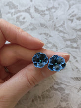 Load image into Gallery viewer, Blue and black flower shaped earrings with blue metallic centers held between finger and thumb. 
