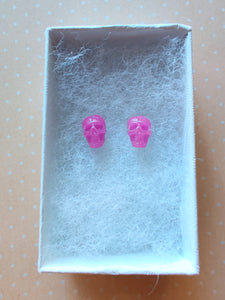 A pair of pink skull shaped earrings in a white paper jewelry box. 