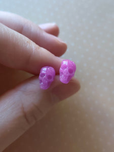 A pair of pink skull shaped earrings held between finger and thumb. 
