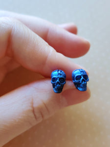 A close of picture of metallic  sapphire colored skull earrings held between finger and thumb. 