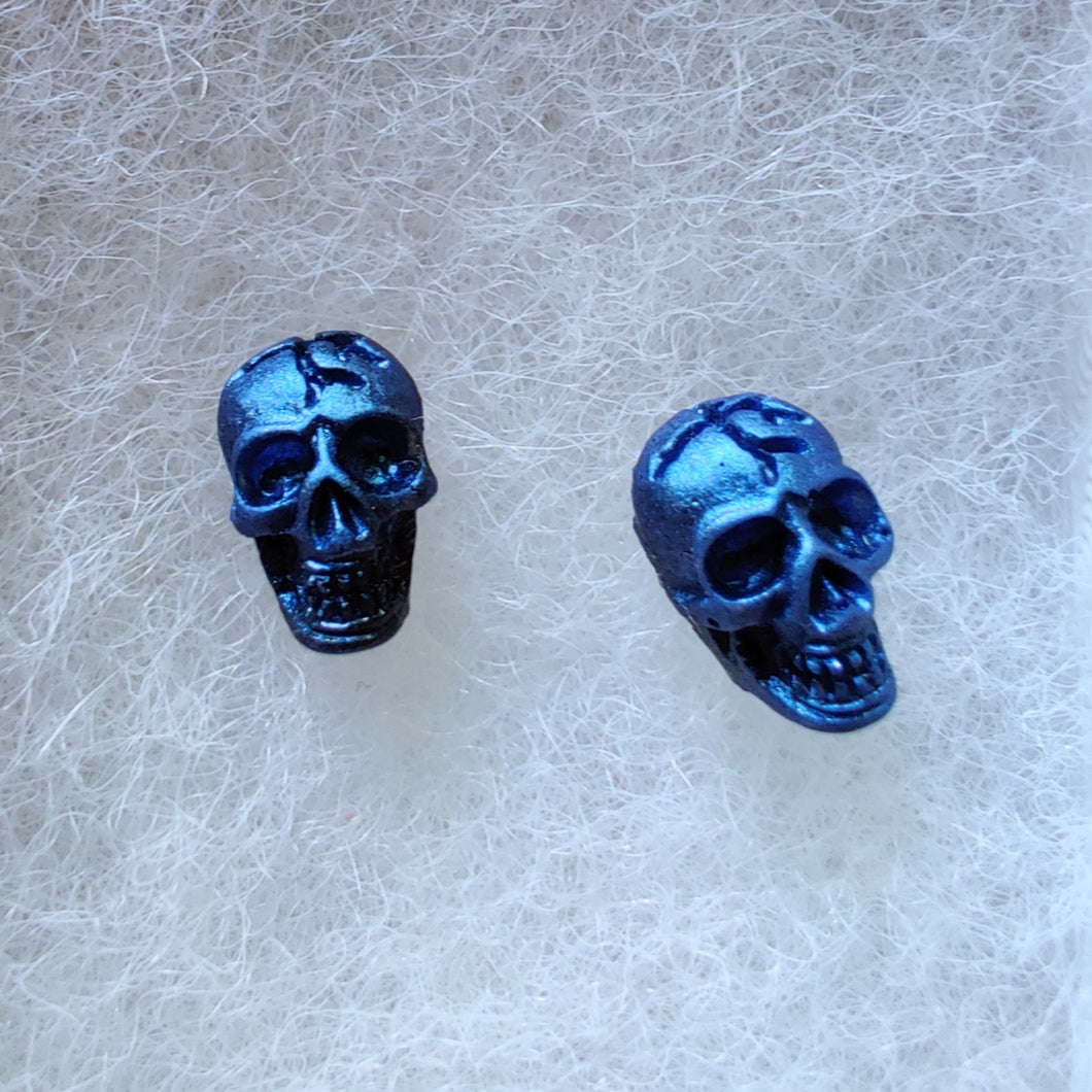 A close of picture of metallic  sapphire colored skull earrings attached to plastic posts. 