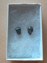 Load image into Gallery viewer, Grey colored skull stud earrings that shimmer silvery in the light inside a white paper jewelry box.  
