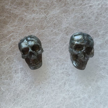 Load image into Gallery viewer, A close of of grey colored skull stud earrings that shimmer silvery in the light. 
