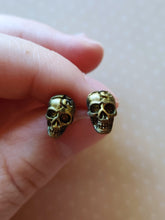 Load image into Gallery viewer, Faux aged gold skull earrings attached to plastic posts, held between finger and thumb.
