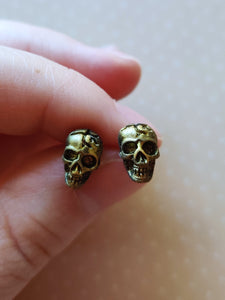 Faux aged gold skull earrings attached to plastic posts, held between finger and thumb.
