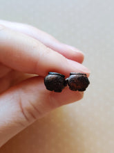 Load image into Gallery viewer, Two miniature flat backed cauldron earrings in black, brushed with a bronze colored metallic pigment. Earrings are held between finger and thumb.
