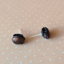 Load image into Gallery viewer, Two miniature flat backed cauldron earrings in black, brushed with a bronze colored metallic pigment. Earrings are displayed on a pale orange background with white polka dots.
