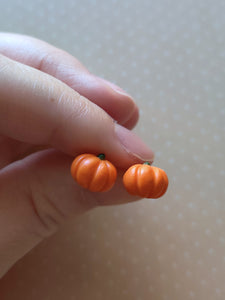 A pair of flat backed, side view, orange pumpkin earrings held between finger and thumb. 