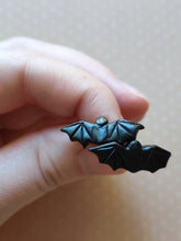 Load image into Gallery viewer, One pair of flat backed bat earrings with relief sculpted detail. Stud earrings are displayed between finger and thumb.
