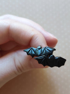 One pair of flat backed bat earrings with relief sculpted detail. Stud earrings are displayed between finger and thumb.