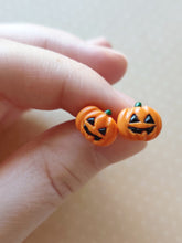Load image into Gallery viewer, A pair of flat backed jack o&#39; lantern earrings with relief sculpted detail and painted on face and stem. The earrings are held between finger and thumb. 
