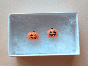 A pair of flat backed jack o' lantern earrings with relief sculpted detail and painted on face and stem. The earrings are inside a white paper jewelry box.