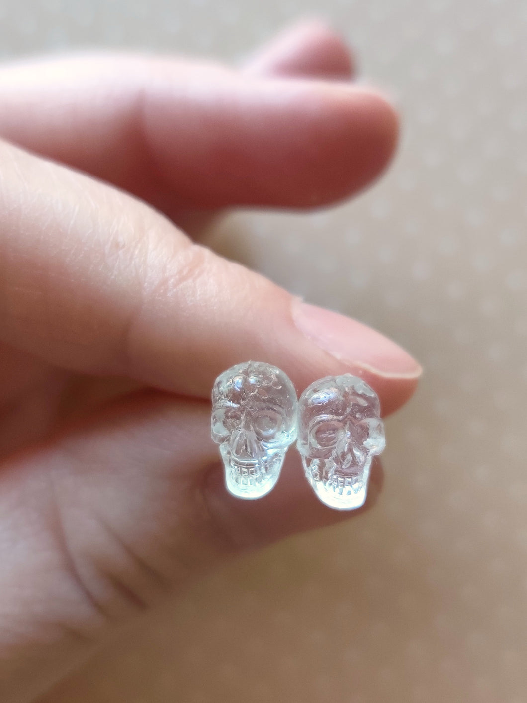 One pair of clear skull stud earrings held between finger and thumb.