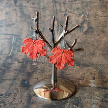 Load image into Gallery viewer, A pair of red maple leaf shaped polymer clay earrings are displayed hanging from a small, polished silver tree shaped display.
