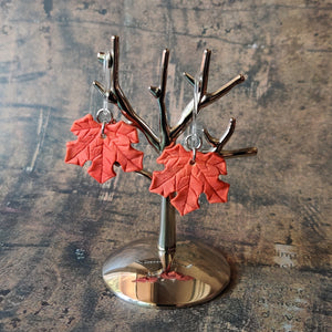 A pair of red maple leaf shaped polymer clay earrings are displayed hanging from a small, polished silver tree shaped display.