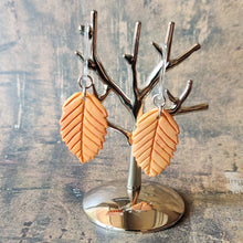 Load image into Gallery viewer, A pair of orange beech leaf shaped polymer clay earrings are displayed hanging from a small, polished silver tree shaped display.
