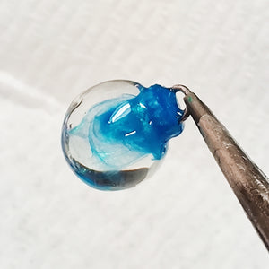a clear miniature Christmas bauble held by the top with small thin pliers. Swirls of a shimmering blue are inside the clear.