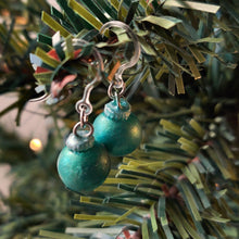 Load image into Gallery viewer, One pair of miniature bauble ornament earrings in a matte finish reflective green. There are clear plastic ear hooks attached by stainless steal jump rings. The earrings are shown hanging from the branch of an artificial Christmas tree. 
