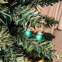 Load image into Gallery viewer, One pair of miniature bauble ornament earrings in a matte finish reflective green. There are clear plastic ear hooks attached by stainless steal jump rings. The earrings are shown hanging from the branch of an artificial Christmas tree.
