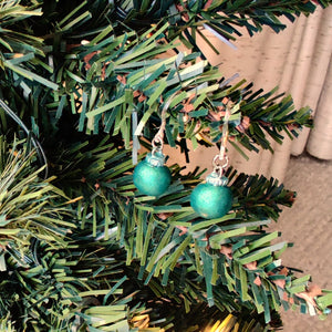 One pair of miniature bauble ornament earrings in a matte finish reflective green. There are clear plastic ear hooks attached by stainless steal jump rings. The earrings are shown hanging from the branch of an artificial Christmas tree.