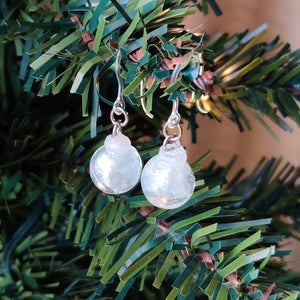 Two miniature Christmas ornament baubles in a clear color with silvery white swirls inside. The earrings are shown hanging from the branch of an artificial Christmas tree. 
