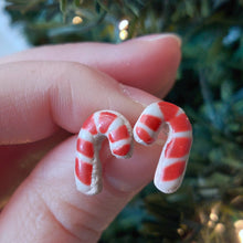 Load image into Gallery viewer, A pair of earrings made to look like sugar cookies decorated as candy canes. The earrings are held between finger and thumb in front of an artificial Christmas tree. 
