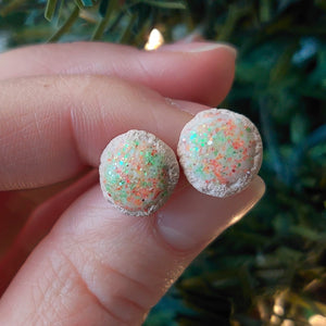 A pair of earrings made to look like sugar cookies decorated with green and red sugar sprinkles. The earrings are held between finger and thumb in front of an artificial Christmas tree. 