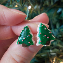 Load image into Gallery viewer, A pair of earrings made to look like sugar cookies decorated like a green Christmas tree with red sugar pearls as ornaments. The earrings are held between finger and thumb in front of an artificial Christmas tree. 
