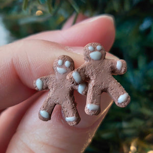 A pair of earrings made to look like gingerbread men. The earrings are held between finger and thumb in front of an artificial Christmas tree. 