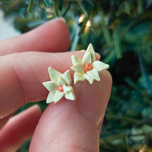 A pair of white Poinsettia earrings held between finger and thumb in front of an artificial Christmas tree.