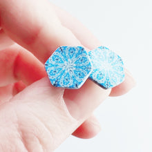 Load image into Gallery viewer, Two hexagon shaped earrings with intricate white, silver and blue patterns held between finger and thumb.
