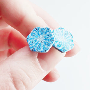 Two hexagon shaped earrings with intricate white, silver and blue patterns held between finger and thumb.