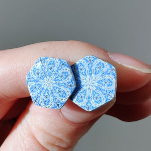 Two hexagon shaped earrings with intricate white, silver and blue patterns held between finger and thumb in the bright sunshine. Small silver sparkles reflect off of the silver areas. 