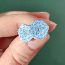 Load image into Gallery viewer, Two hexagon shaped earrings with intricate white, silver and blue patterns held between finger and thumb in bright sunlight.
