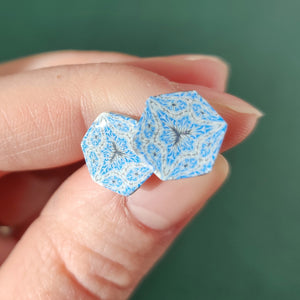 Two hexagon shaped earrings with intricate white, silver and blue patterns held between finger and thumb in bright sunlight.