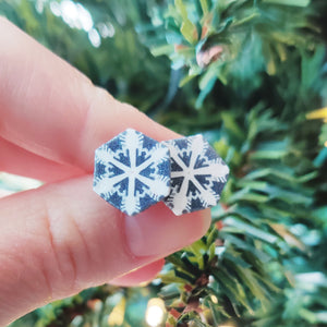 Two hexagon shaped earrings with a white and silver snowflake design held between finger and thumb before an artificial Christmas tree branch. 