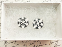 Load image into Gallery viewer, Two hexagon shaped earrings with a white and silver snowflake design inside a white paper jewelry box. 
