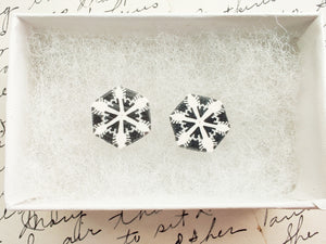 Two hexagon shaped earrings with a white and silver snowflake design inside a white paper jewelry box. 